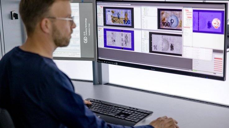 A person is working at a desk with two large computer monitors displaying images of currency notes