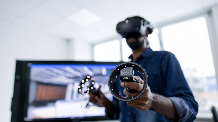 A professional using a virtual reality headset and controllers, standing in an office environment with a monitor displaying a virtual simulation in the background.