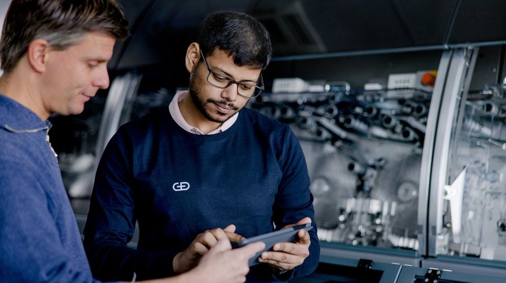 Two professionals in a workshop discussing over a digital tablet, with industrial machinery in the background.