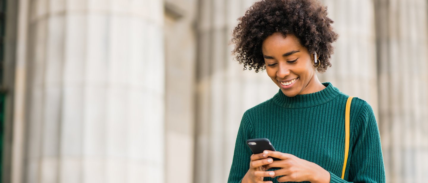 Person in a green sweater standing outside in front of a large pillar and using a smartphone.