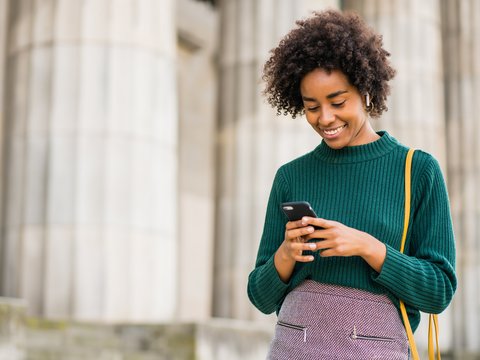  Person in a green sweater standing outside in front of a large pillar and using a smartphone.