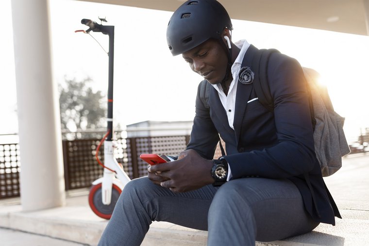 Young businessman with scooter sitting on stairs at sunset using mobile phone and earphones