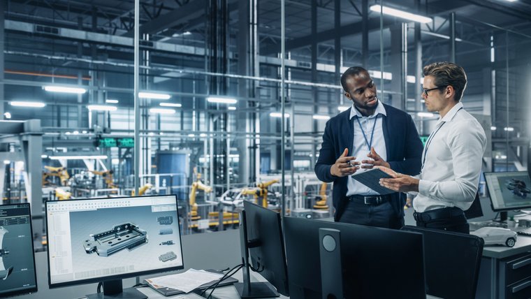 Two people discussing, in the background computer monitors 3D designs and robotic machines