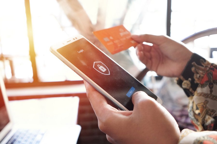 A left hand holds a smartphone verifying a payment on a login screen, the right hand holds a credit card.