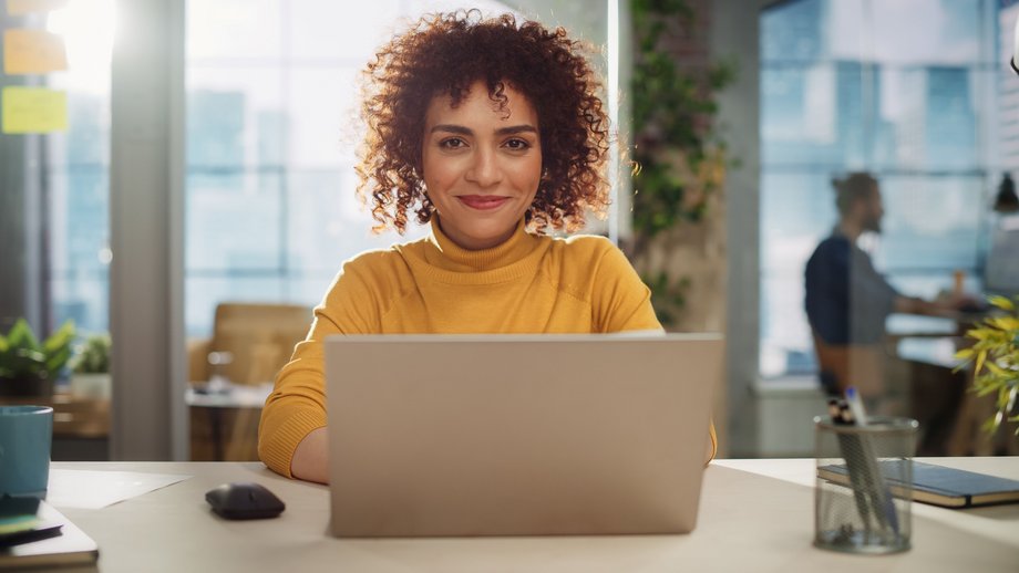 A person sitting in an office with a notebook with two other peoples chatting in the background