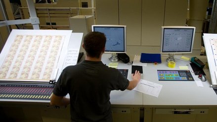 A man stands in front of a sheet of banknotes, next to it are two monitors on a desk