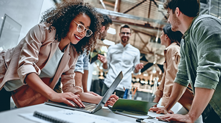 Some people stand together leaning on a desk, a woman operates a laptop