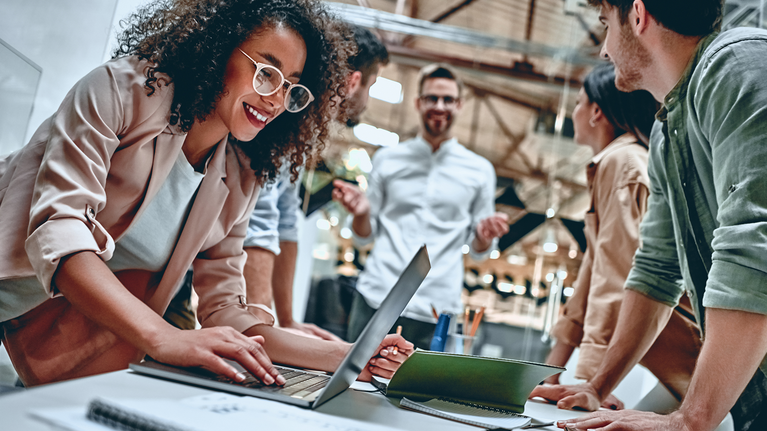 Some people stand together leaning on a desk, a woman operates a laptop